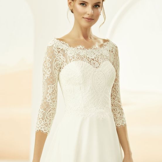 Eine Frau in einem weißen Hochzeitskleid mit langen Spitzenärmeln posiert drinnen und lächelt in die Kamera.