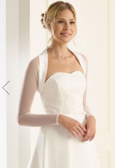 Eine Frau trägt ein weißes Hochzeitskleid mit langen Ärmeln und einer Halskette. Sie steht vor einer weißen Wand.