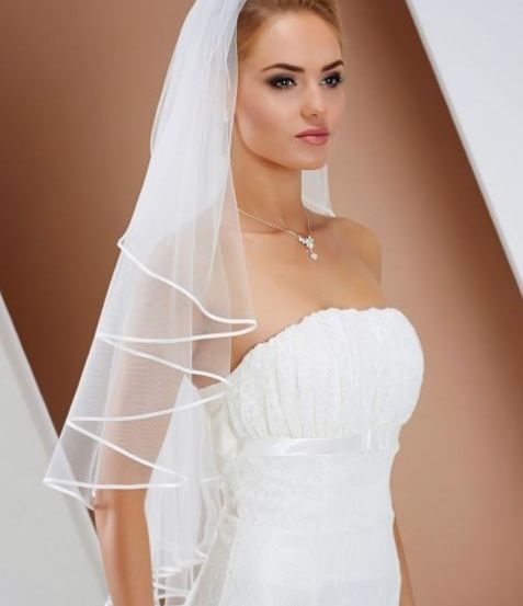 Eine Frau in einem weißen Hochzeitskleid mit Schleier posiert vor einem beigen Hintergrund und trägt eine Halskette mit Anhänger.