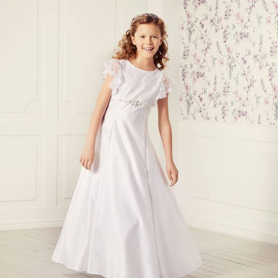 Ein junges Mädchen in einem weißen Kleid mit Spitzenärmeln und einem Perlengürtelst steht in einem Raum mit Blumentapeten und weißen Wänden.