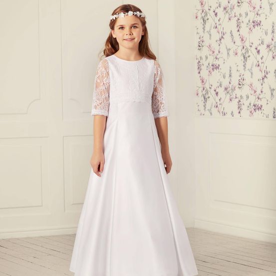 Ein junges Mädchen steht in einem Raum und trägt ein weißes Kleid mit Spitzenärmeln und einem Blumenkopfband. Sie lächelt und posiert für ein Foto.