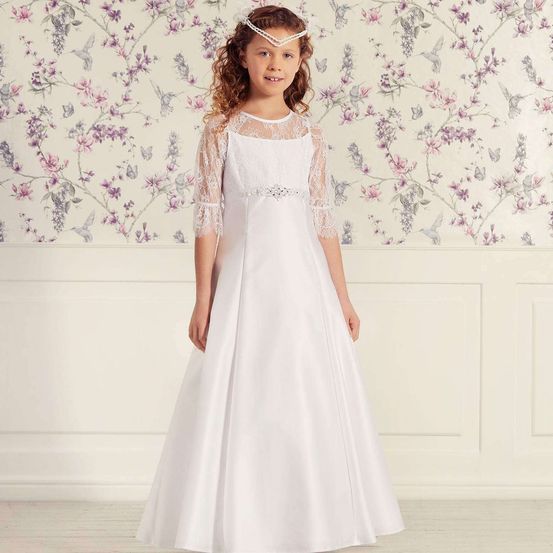 Ein kleines Mädchen in einem weißen Kleid mit Spitzenärmeln steht vor einer floralen Tapete.