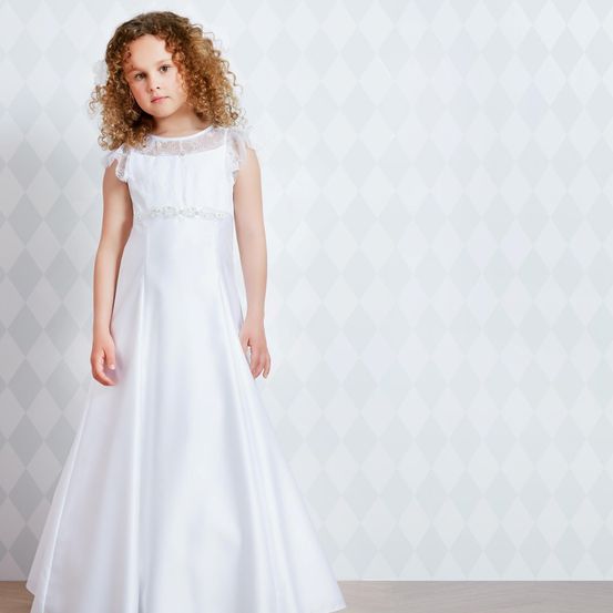 Ein kleines Mädchen mit lockigen Haaren steht vor einer rautenförmigen Wand und trägt ein weißes, langärmeliges Kleid mit Spitze und Silberbesatz.