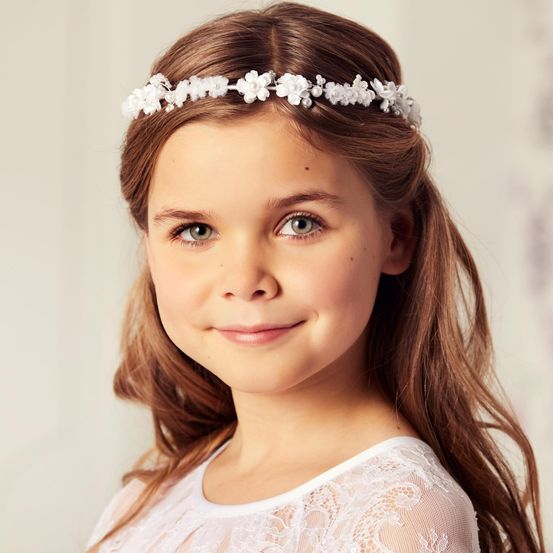 Ein junges Mädchen mit braunen Haaren, das eine weiße Blumenkopfband und ein weißes Spitzenkleid trägt, lächelt in die Kamera.