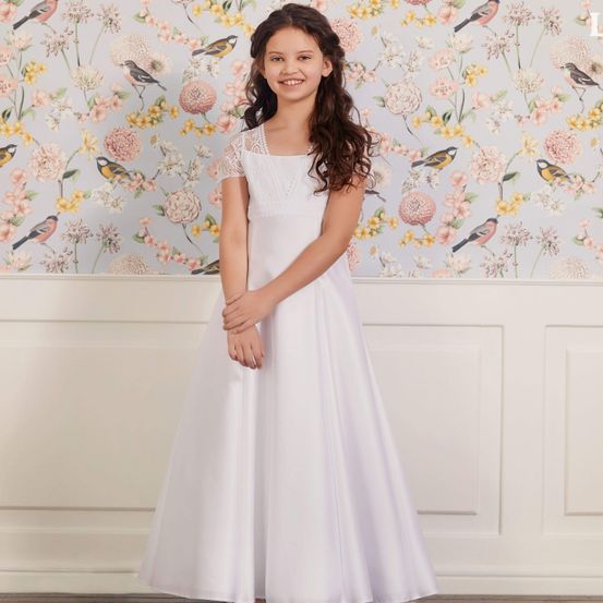 Ein junges Mädchen mit lockigen Haaren, das ein weißes Kleid mit Spitzenärmeln trägt, steht vor einem floralen Tapetenhintergrund.