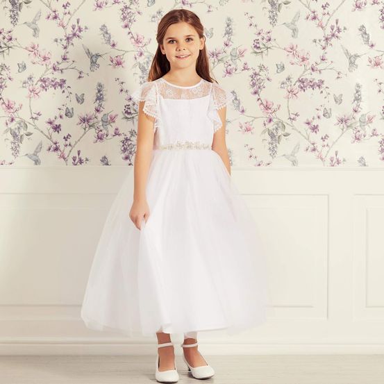 Ein kleines Mädchen in einem weißen Kleid und weißen Schuhen steht vor einer Blumentapete. Sie hat braune Haare und schaut zur Seite.