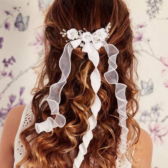 Eine Frau mit lockigem Haar trägt ein weißes Blumenhaaraccessoire. Das Accessoire hat fließende Bänder. Der Hintergrund ist Blumentapete.