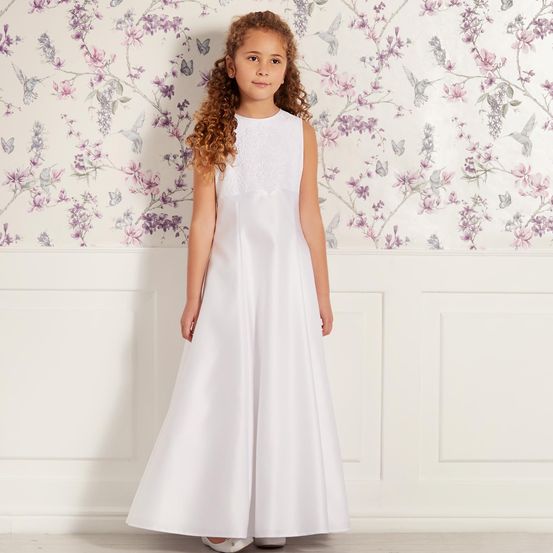 Ein kleines Mädchen mit lockigem Haar steht vor einer weißen Wand mit Blumentapete und trägt ein weißes ärmelloses Kleid.