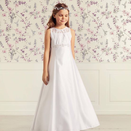Ein junges Mädchen steht vor einer Wand mit Blumentapete und trägt ein weißes Kleid mit Spitzendetails und einer Blumenhaarband.