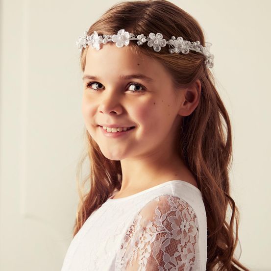 Ein junges Mädchen mit langen braunen Haaren lächelt und trägt ein weißes Kleid und eine Kopfband mit weißen Blumen und Kristallen.