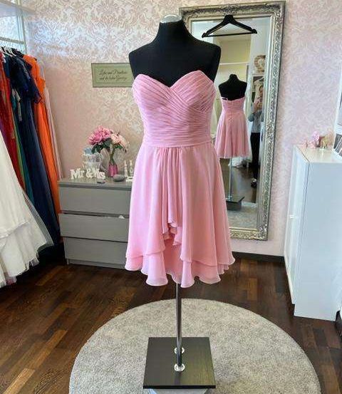 Ein rosa Brautjungfernkleid an einem Mannequin, mit einem Spiegel, einer Kommode und einem Regal mit Kleidung im Hintergrund.