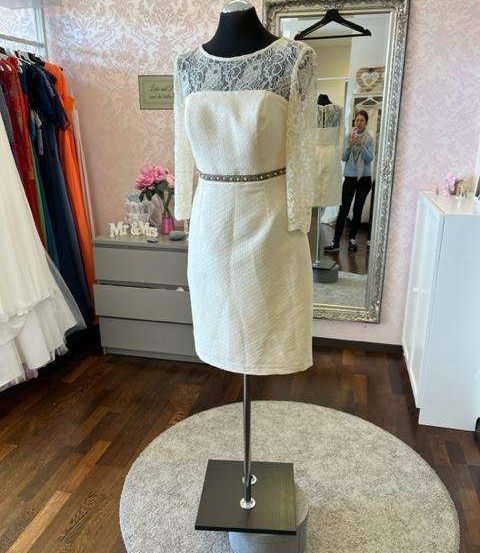 Ein weißes Kleid mit Spitzenärmeln und einem bestickten Gürtel wird auf einem Mannequin in einem rosa Raum mit Holzboden ausgestellt.