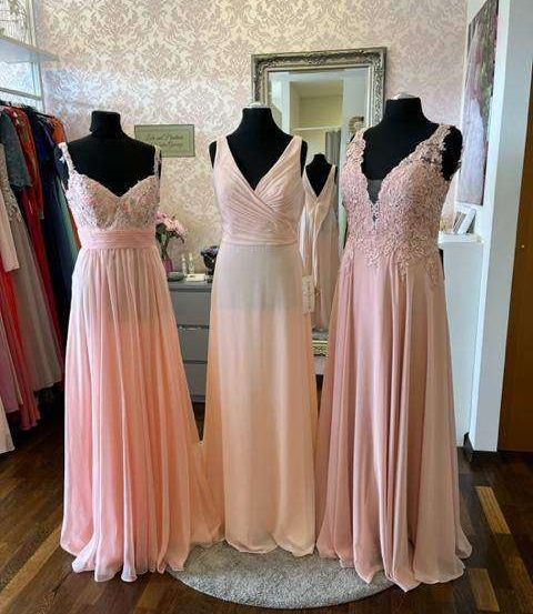 Drei rosa Kleider sind auf Mannequins in einem Boutique ausgestellt. Die Kleider haben aufwendige Spitzendesigns und einen V-Ausschnitt. Die Kleider stehen auf einem Teppich auf einem Holzboden.