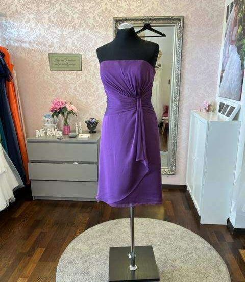 Ein lila Cocktailkleid auf einem Mannequin in einem Raum mit rosa Wand und Spiegel, zusammen mit einem Schild, das Mr & Mrs steht.