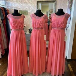 Drei Mannequins in rosa Kleidern mit silbernen Gürteln stehen auf einem runden Teppich in einem Boutique. Dahinter befindet sich ein Spiegel, der weitere Kleider reflektiert, und eine Wand mit einem Schild.