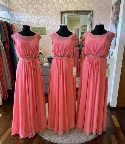 Drei Mannequins in rosa Kleidern mit silbernen Gürteln stehen auf einem runden Teppich in einem Boutique. Dahinter befindet sich ein Spiegel, der weitere Kleider reflektiert, und eine Wand mit einem Schild.