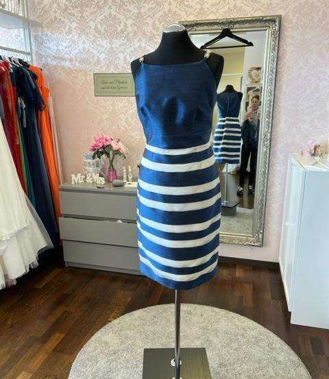 Ein blau-weiß gestreiftes Kleid auf einem Mannequin wird in einem Raum mit verspiegelter Wand und einem Kleiderständer ausgestellt.