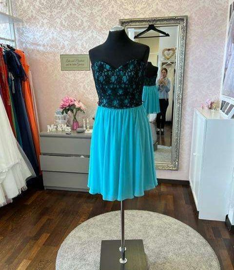 Ein Mannequin zeigt ein türkisfarbenes und schwarzes Kleid in einem Raum mit rosa Tapete, einem Kommode und einem Spiegel. Eine Person spiegelt sich im Spiegel.