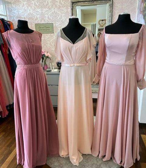 Drei rosa Kleider, die auf Mannequins in einem Raum mit rosa Wand und Spiegel ausgestellt sind. Ein Schild mit der Aufschrift 'Leben und Freude'.