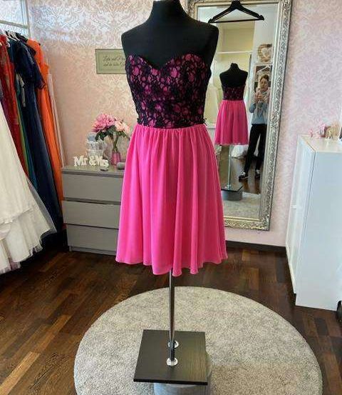 Ein Dummy zeigt ein pinkes und schwarzes Kleid in einem Boutique. Das Kleid hat einen schwarzen Spitzenoberteil mit pinken Blumen. Dahinter reflektiert ein Spiegel einen anderen Dummy und eine Person, die ein Foto macht.