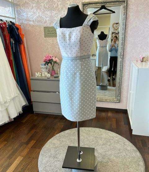 Ein weißes Kleid mit Punkten auf einem Mannequin wird in einem Raum mit einer rosa Wand ausgestellt. Ein Spiegel und ein Schrank befinden sich hinter dem Kleid.