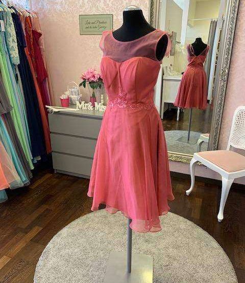 Ein Dummy zeigt ein rosa Cocktailkleid in einer Umkleidekabine. Ein Spiegel reflektiert den Raum und einen Schrank mit einer Blumenvase. Stühle und Kleiderständer sind ebenfalls sichtbar.