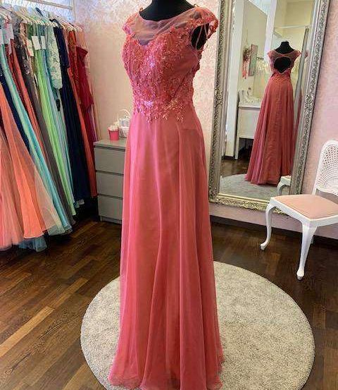 Ein Mannequin präsentiert ein korallenfarbenes Abendkleid auf einem runden Teppich. Das Kleid ist mit Spitze und Pailletten verziert. Dahinter reflektiert ein Spiegel das Kleid und einen rosa Stuhl. Links steht ein Kleiderständer mit verschiedenen farbigen Kleidern.