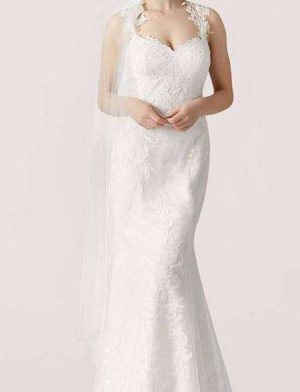 Eine Frau in einem weißen Hochzeitskleid mit Spitzendetails posiert mit den Händen auf der Taille. Sie trägt einen Schleier.