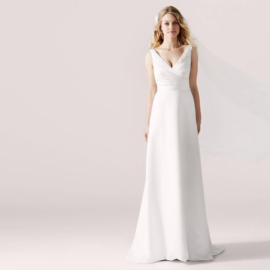 Eine blonde Frau steht und trägt ein weißes Hochzeitskleid und einen Schleier vor einem hellrosa Hintergrund.