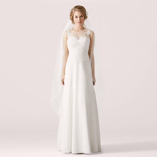 Eine Frau in einem weißen Hochzeitskleid mit Spitzendetails und einem Schleier steht vor einem hellen Hintergrund.