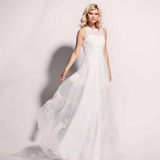 Eine Frau mit blonden Haaren trägt ein weißes Hochzeitskleid mit Spitzendetails vor einem weißen Hintergrund.