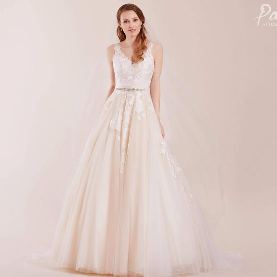 Eine Braut steht in einem cremefarbenen Hochzeitskleid mit Spitzendetails und einem silbernen Gürtel und posiert vor einem hellrosa Hintergrund.