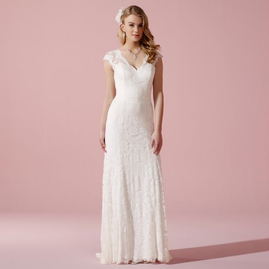 Eine Frau steht vor einem rosa Hintergrund und trägt ein weißes Hochzeitskleid mit Spitze und einer Blume im Haar.