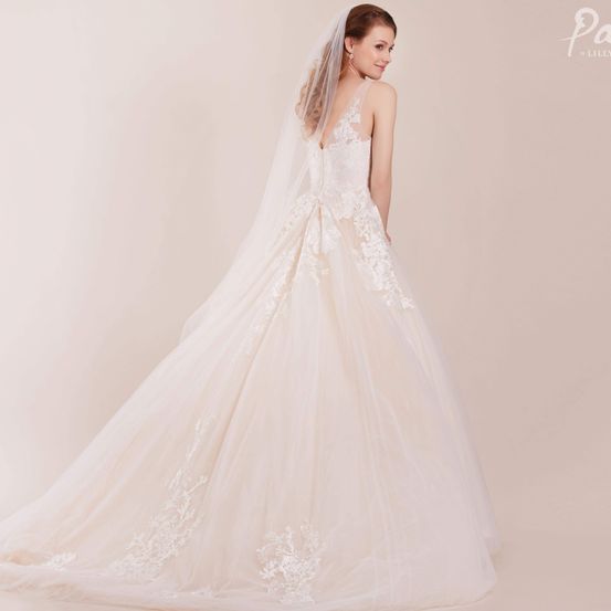 Eine Braut in einem weißen Hochzeitskleid mit aufwendiger Spitze und einem langen Schleier, vor einem hellrosa Hintergrund stehend.