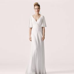 Eine Frau in einem weißen Kleid mit gerüschten Ärmeln steht vor einem hellrosa Hintergrund.