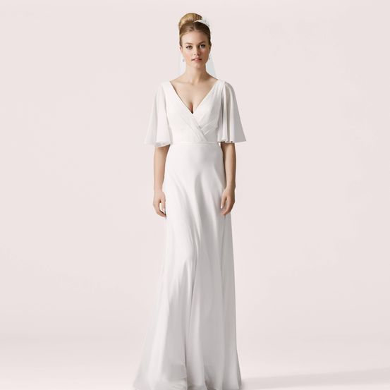 Eine Frau in einem weißen Kleid mit gerüschten Ärmeln steht vor einem hellrosa Hintergrund.