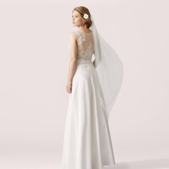 Eine Braut in einem weißen Kleid mit einem verzierten Spitzenrücken und Schleier posiert vor weißem Hintergrund.