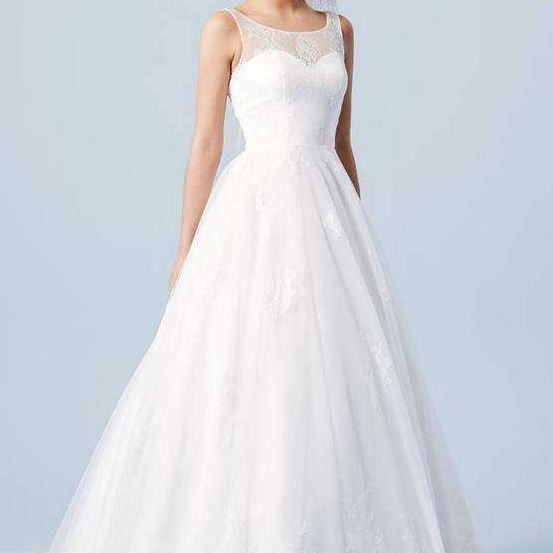 Eine Frau in einem weißen Hochzeitskleid mit Spitzendetails posiert für ein Foto vor blauem Hintergrund.