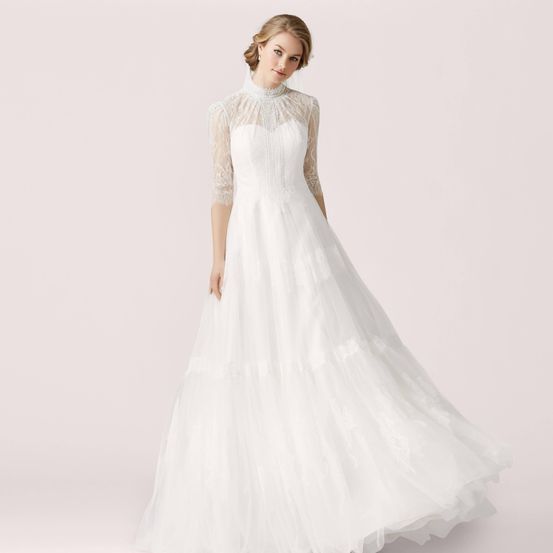 Eine Frau in einem weißen Hochzeitskleid mit Spitzenärmeln und hohem Kragen steht vor einem rosa Hintergrund.