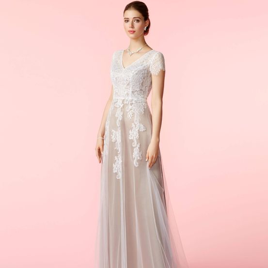 Eine Frau in einem weißen und grauen Kleid mit Spitzendetails steht vor einem rosa Hintergrund. Sie trägt eine Perlenkette und Ohrringe.