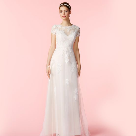 Eine Frau in einem weißen Hochzeitskleid mit Spitzendetails steht vor einem rosa Hintergrund. Sie trägt eine Kopfband und Ohrringe.