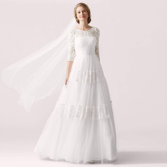 Eine Braut steht in einem rosa Raum, trägt ein weißes Hochzeitskleid mit Spitzendetails, einen Schleier und lächelt für ein Foto.