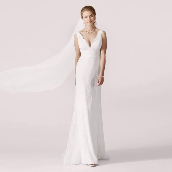 Eine Frau steht und trägt ein weißes Hochzeitskleid aus Spitze mit V-Ausschnitt und Schleier. Sie hat ihr Haar hochgebunden und trägt eine Halskette und Ohrringe.