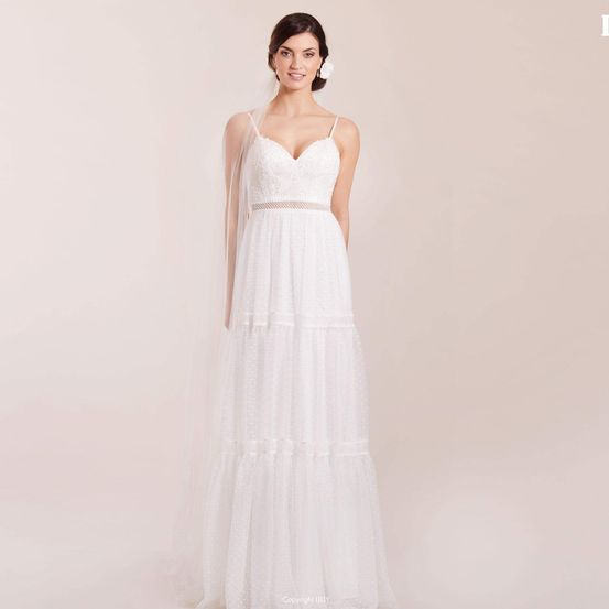 Eine Frau in einem weißen Hochzeitskleid mit einer mehrschichtigen Rock und einem Schleier steht vor einem rosa Hintergrund und lächelt.