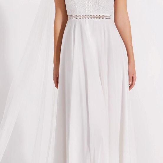 Eine Frau trägt ein weißes, mit Spitze verziertes Kleid mit wehendem Rock und Schleier, vor weißem Hintergrund stehend.