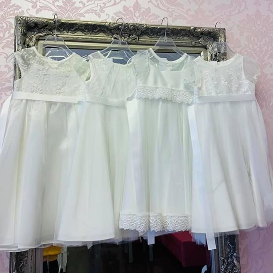 Drei weiße Kleider hängen an einem Kleiderbügel vor einem Spiegel. Die Kleider haben Spitzendetails und sind wahrscheinlich für einen besonderen Anlass.