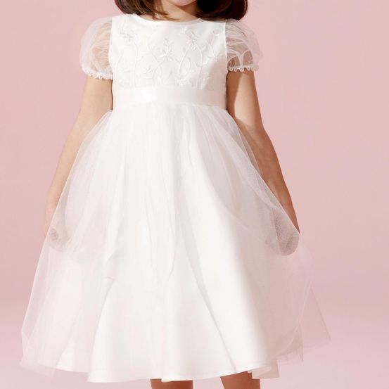 Ein junges Mädchen in einem weißen Kleid mit Spitzendetails steht vor einem rosa Hintergrund. Das Kleid hat einen taillierten Oberteil und einen vollen Rock.