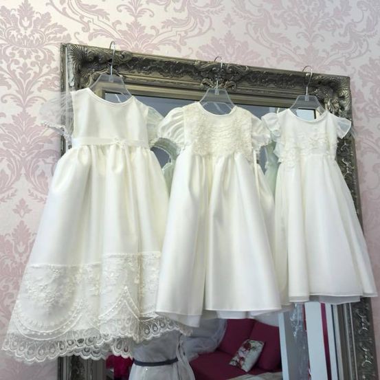 Drei weiße Kleider mit Spitzendetails hängen an einem Spiegel mit Blumentapete. Ein roter Couch mit Kissen spiegelt sich im Spiegel.