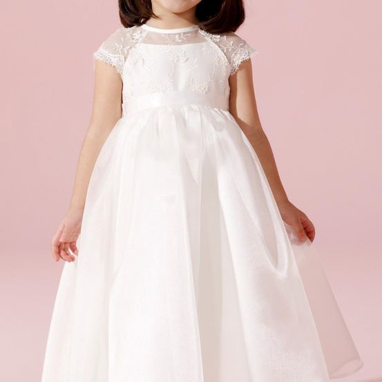 Ein junges Mädchen trägt ein weißes Kleid mit Spitzendetails an den Schultern und einem Satin-Finish. Sie steht vor einem rosa Hintergrund.