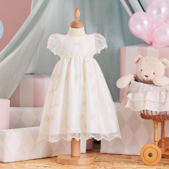 Ein weißes Kleid mit Spitzenbesatz steht auf einem Mannequin, umgeben von rosa Luftballons, einem weißen Teddybären und rosa und weißen Kisten.